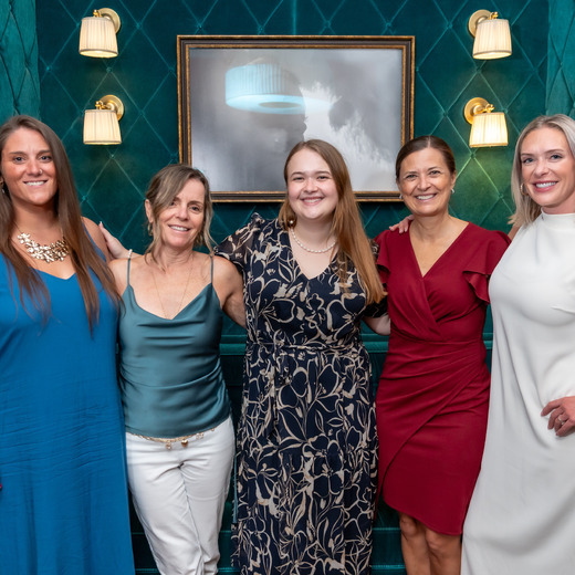 A group of five women in cocktail attire link arms and smile for the camera