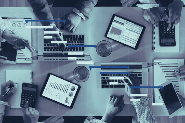 A desk shows multiple tablets and laptops with visual overlays representing the connection of different systems and reports. Multiple hands work on laptops, calculators, and paper across the desk.