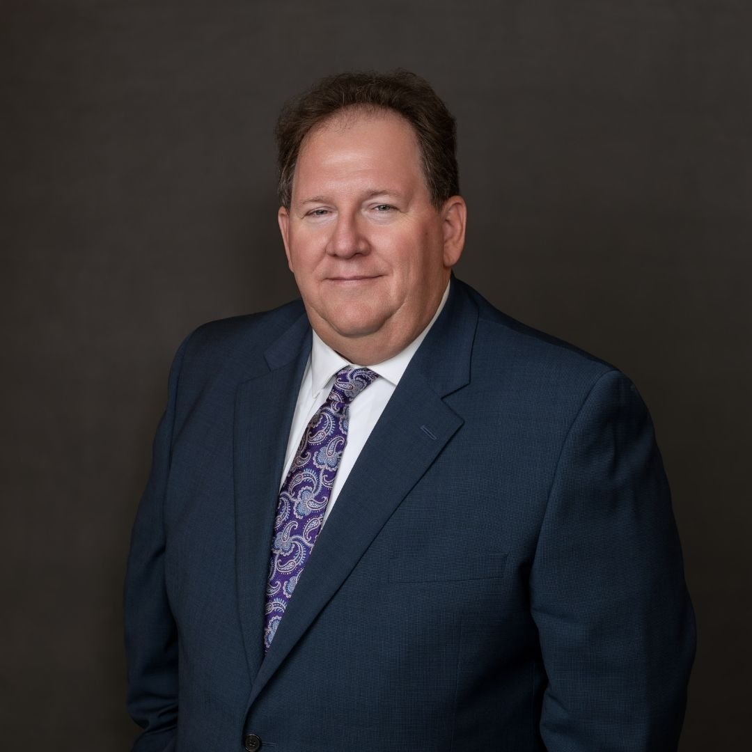 Richard Demoret, Director of Engineering, smiles at the camera in a gray suit and tie against a dark background