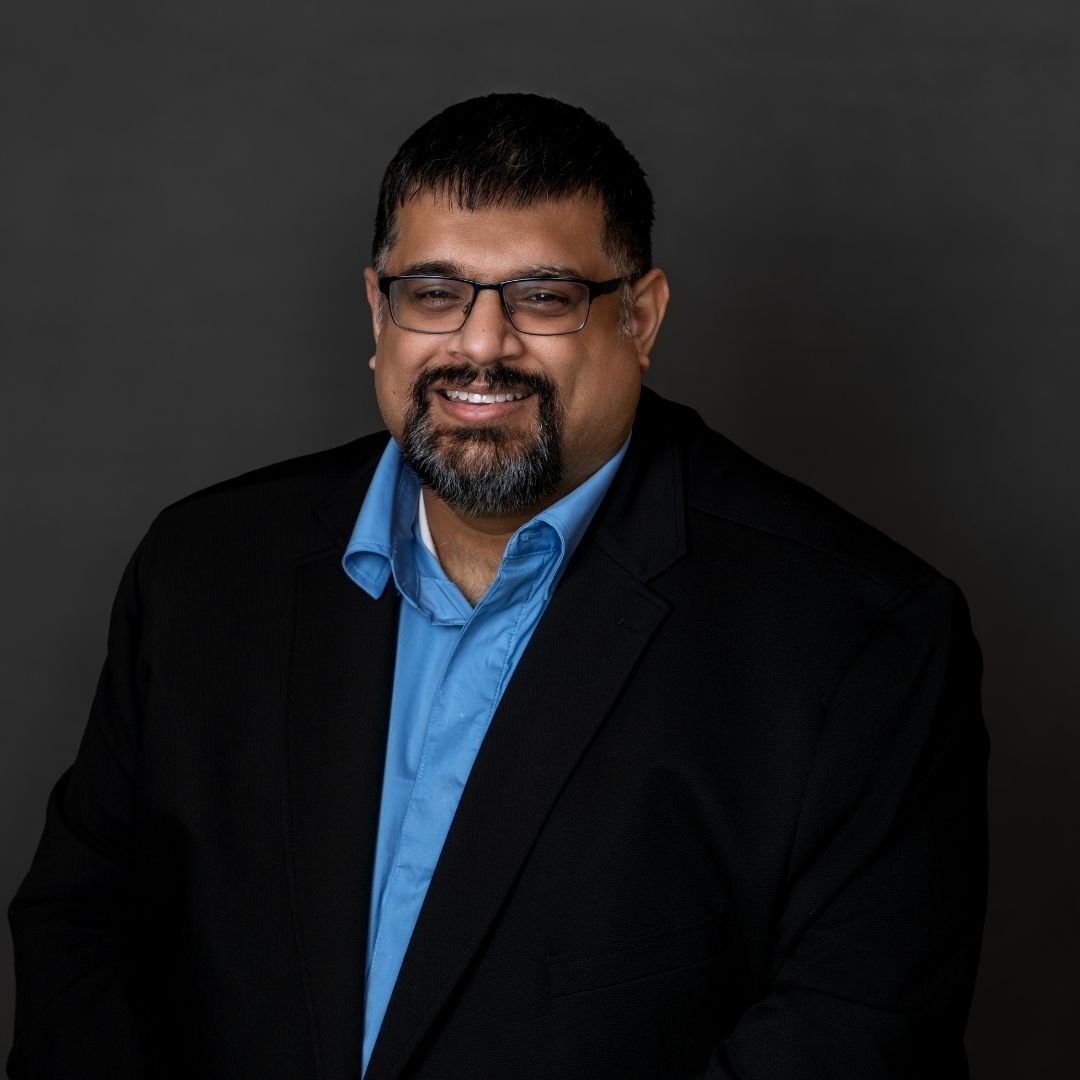 Vinod Kalra, Director of Managed Services, smiles at the camera in a black suit against a dark background